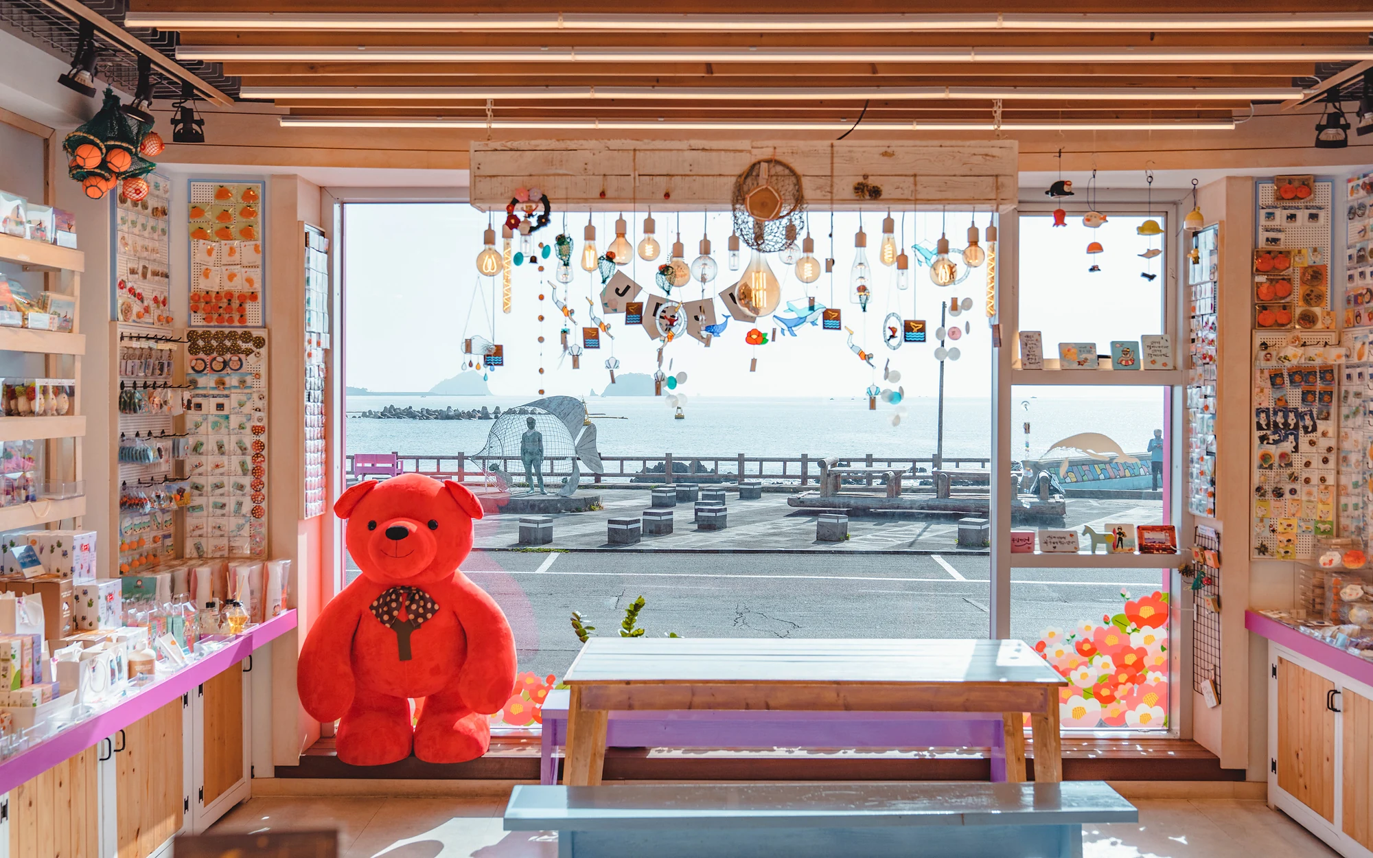 창 너머로 법환포구 바다가 보이는 제스토리 매장 안. 창가에 빨간 곰인형이 앉아 있다